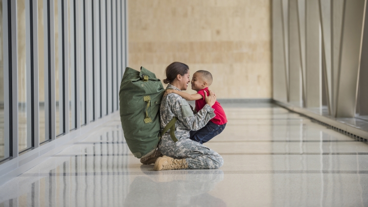 Black female soldier in camouflage with large backpack hugging little boy in red shirt in a hallway