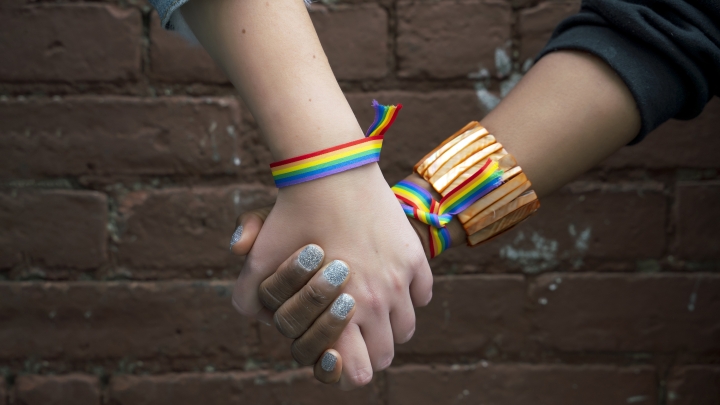 Two hands clasped, one white and one brown with sparkly nail polist, both with rainbow wrist bands