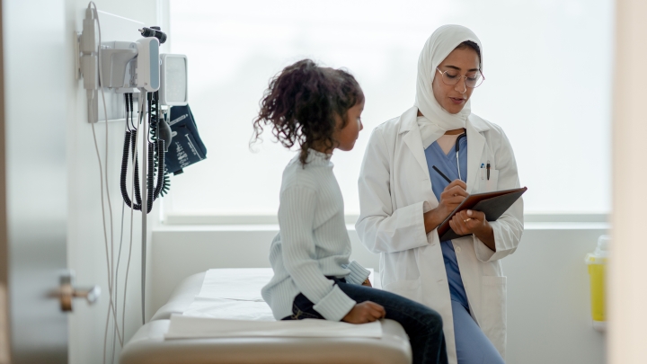 Female doctor wearing glasses and white hajib talking to young girl in exam room