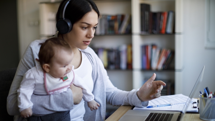 Young mother with dark hair wearing headphones, holding a baby, and looking at a laptop