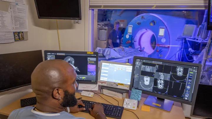 Radiology technolgist looking at scans on a computer monitor. Through the window the CT scanner can be seen.