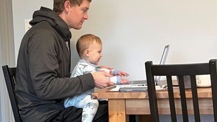 Man sitting at a table working on a laptop with a baby on his lap