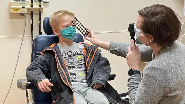 Young blond-haired boy seated in chair wearing jacket and mask getting eye exam