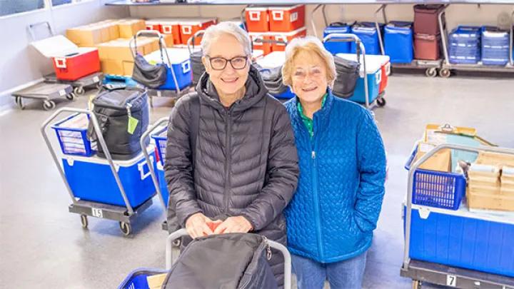 Two women stand together in a warehouse with a shopping cart