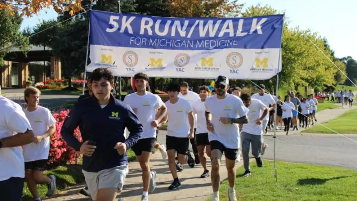 A group of runners participates in a “5K Run/Walk for Michigan Medicine” event, passing under a large event banner on a sunny outdoor path.