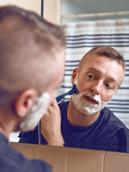 Man shaving in front of mirror with white and blue striped shower curtain in the background