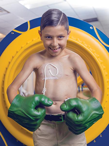 Shirtless boy with hospital tube smiling and wearing large green Hulk gloves 