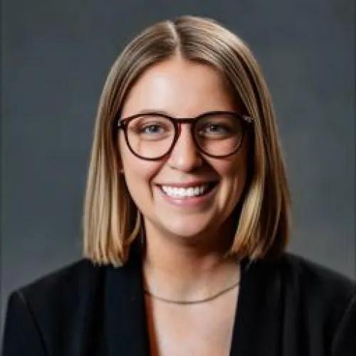 Smiling portrait of Shae Maier. She is wearing glasses, a dark blazer, and a simple necklace.