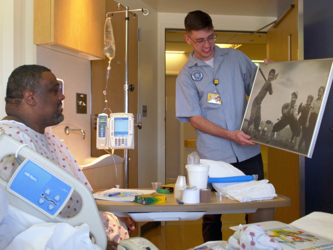 Male volunteer displaying old baseball art poster to older Black male patient in hospital bed