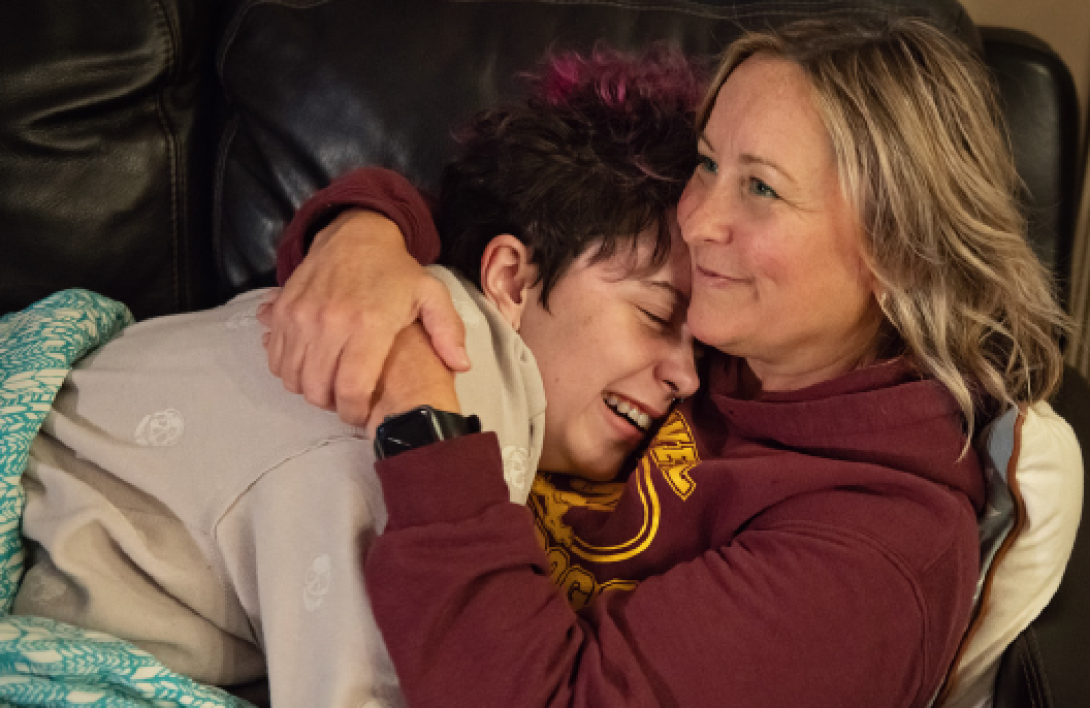 A smiling mom hugs her laughing adolescent son. They are snuggled under a blanket on a couch.