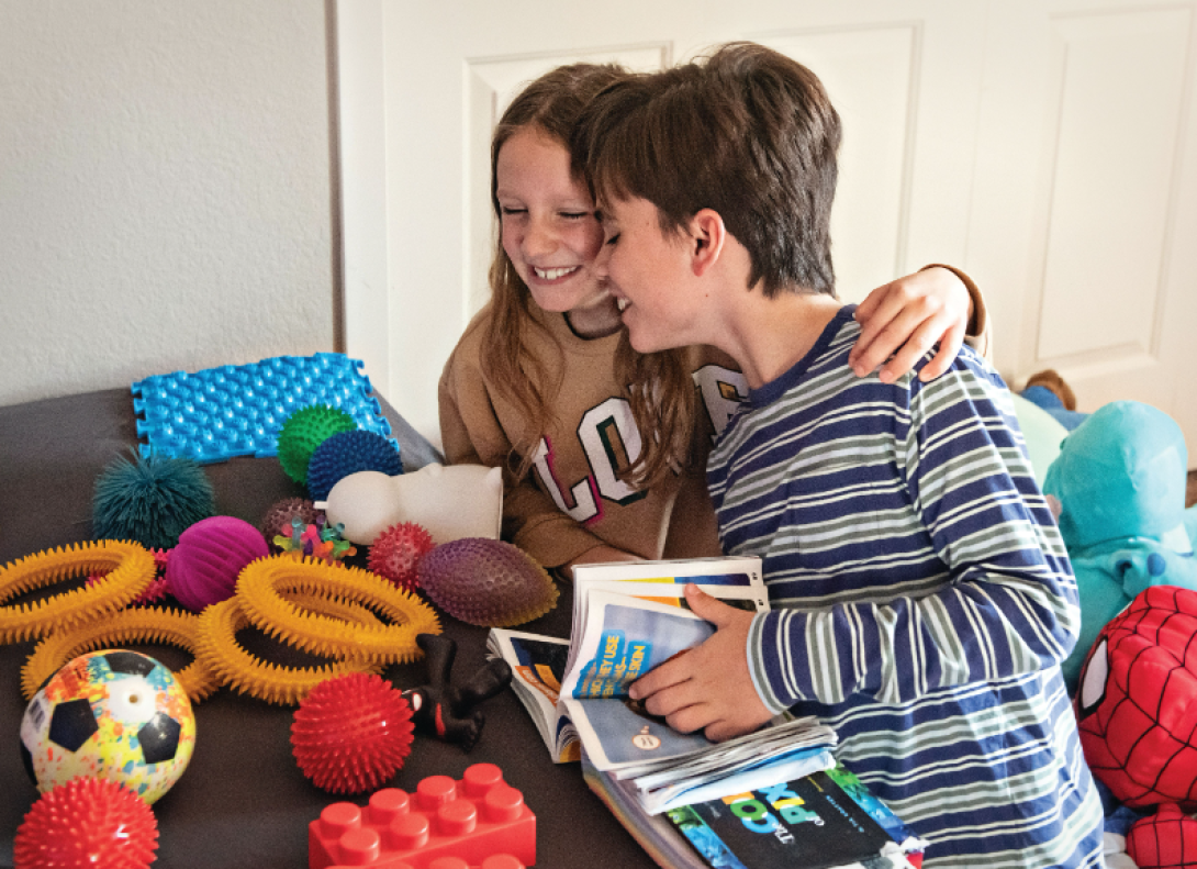 A sister and brother hug each other. They are surrounded by toys.