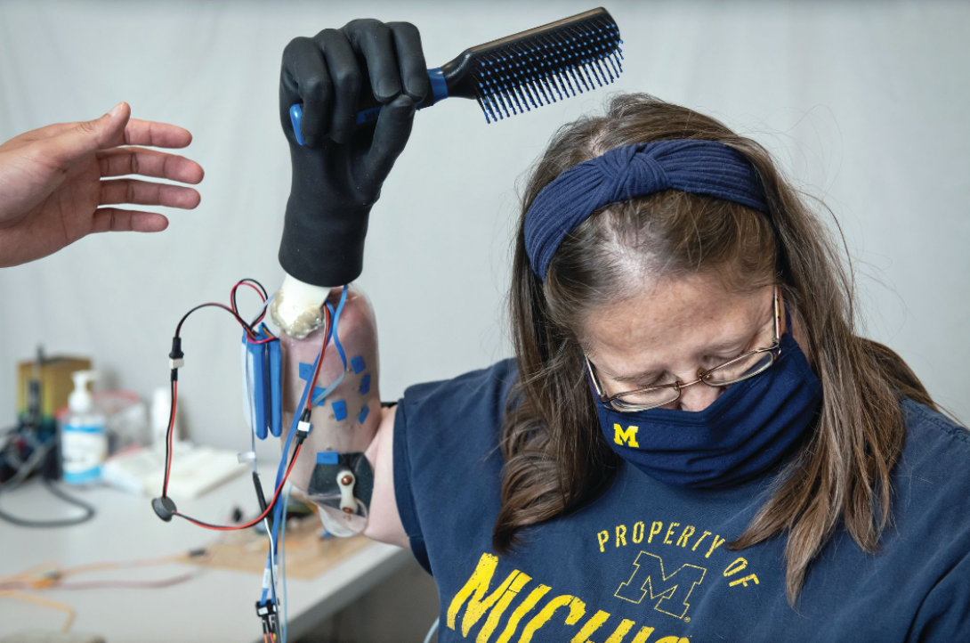 A white woman with glasses who is wearing a U-M T-shirt and U-M face mask brushes her hair using a prosthetic hand