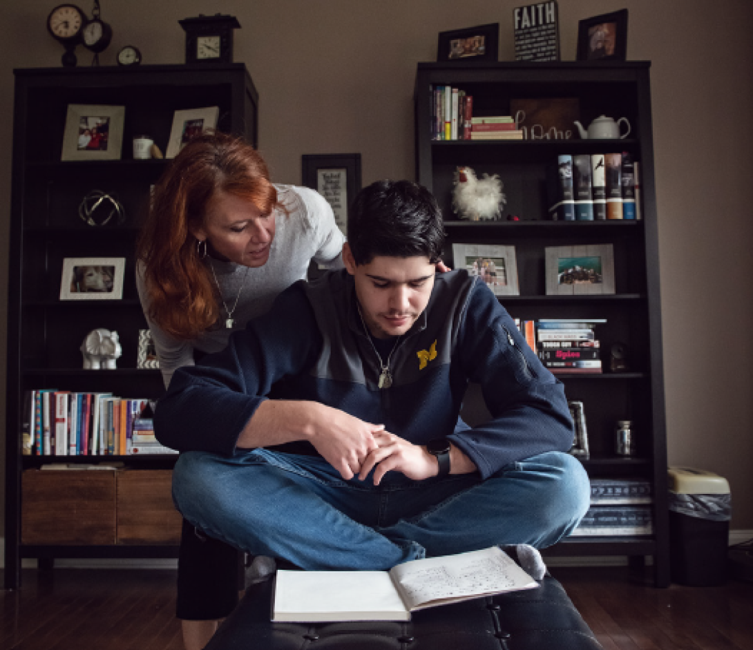 An adolescent boy sits cross legged with a notebook open in front of him. His mom leans over his shoulder to look at the notebook with him. They are in a living room with bookshelves.