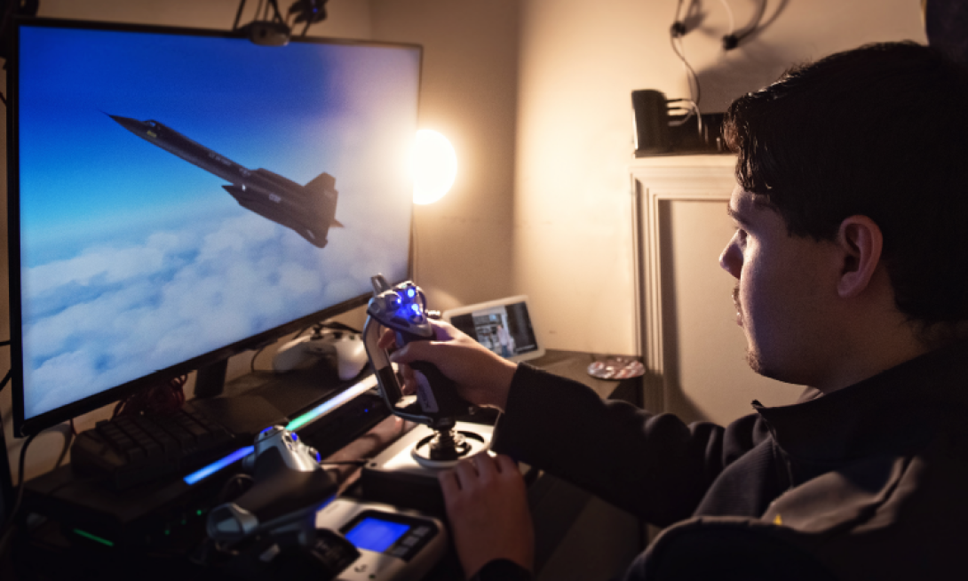 A teen boy sits in front of a large computer monitor, playing a video game. The monitor shows a realistic looking aircraft in a blue sky with clouds.