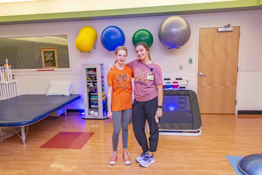 teenage girl in orange shirt and orange sneakers posing smiling with young adult woman in maroon colored shirt in physical therapy gym room