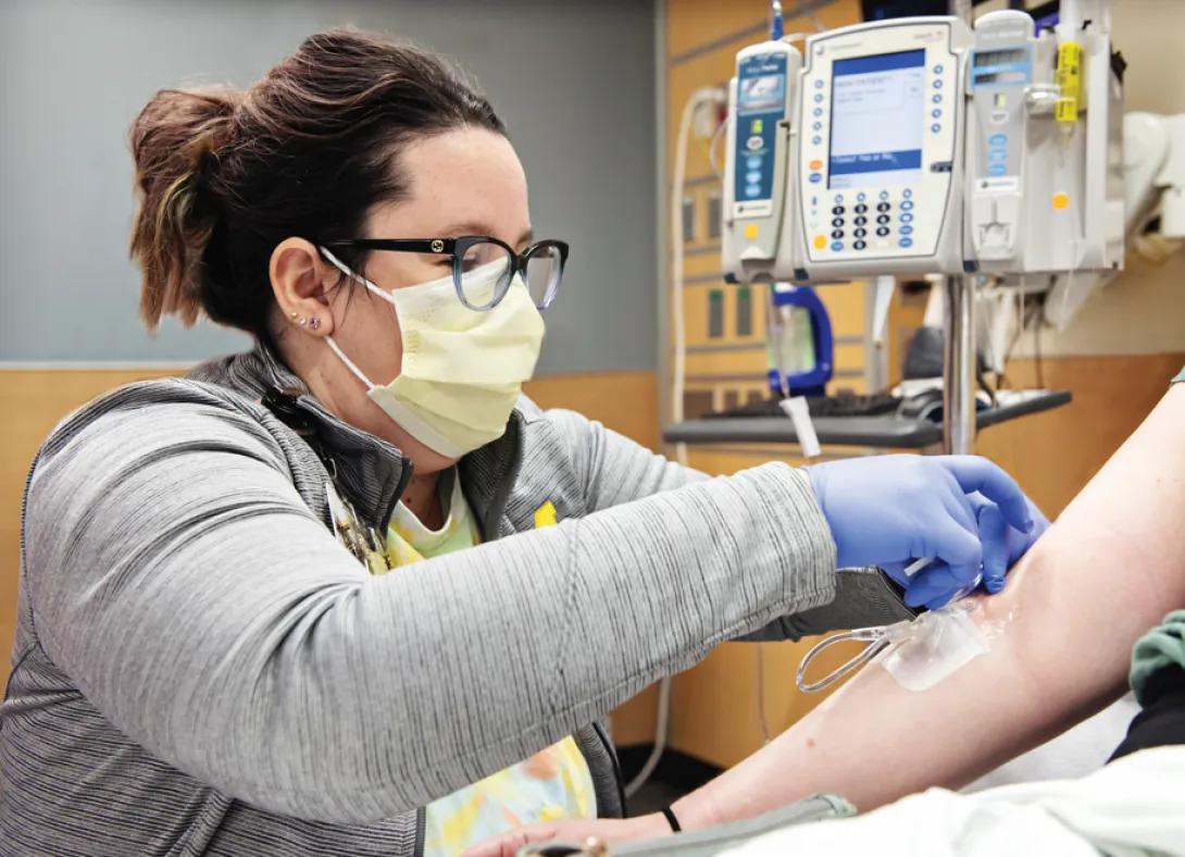 A nurse wearing a mask and gloves attaches an IV to a patient's arm