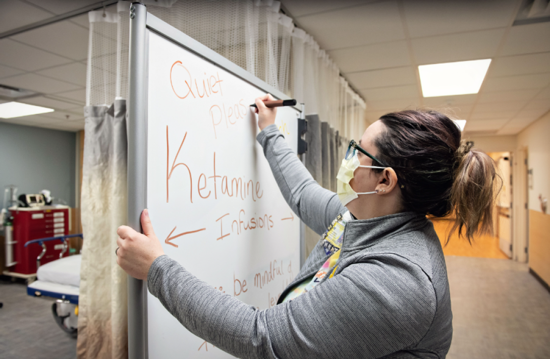 A nurse in a mask writes "Quiet please" on a whiteboard at the entrance of the ketamine infusion bay in the hospital