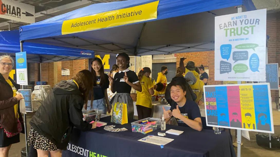 TAC TAC sitting at AHI's booth at the 2022 Youth Summit at the Big House