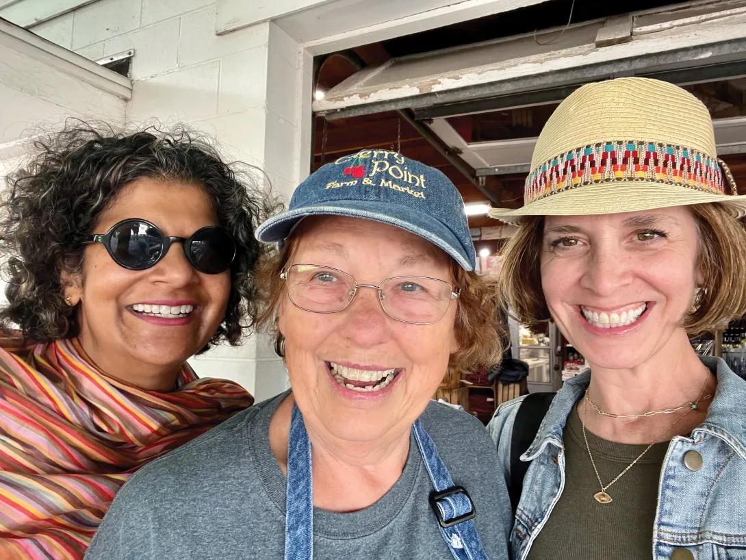Three women smiling at the camera. From the left, one woman is wearing sunglasses and a striped scarf, another woman is wearing a baseball hat, and the third woman is wearing a straw hat.