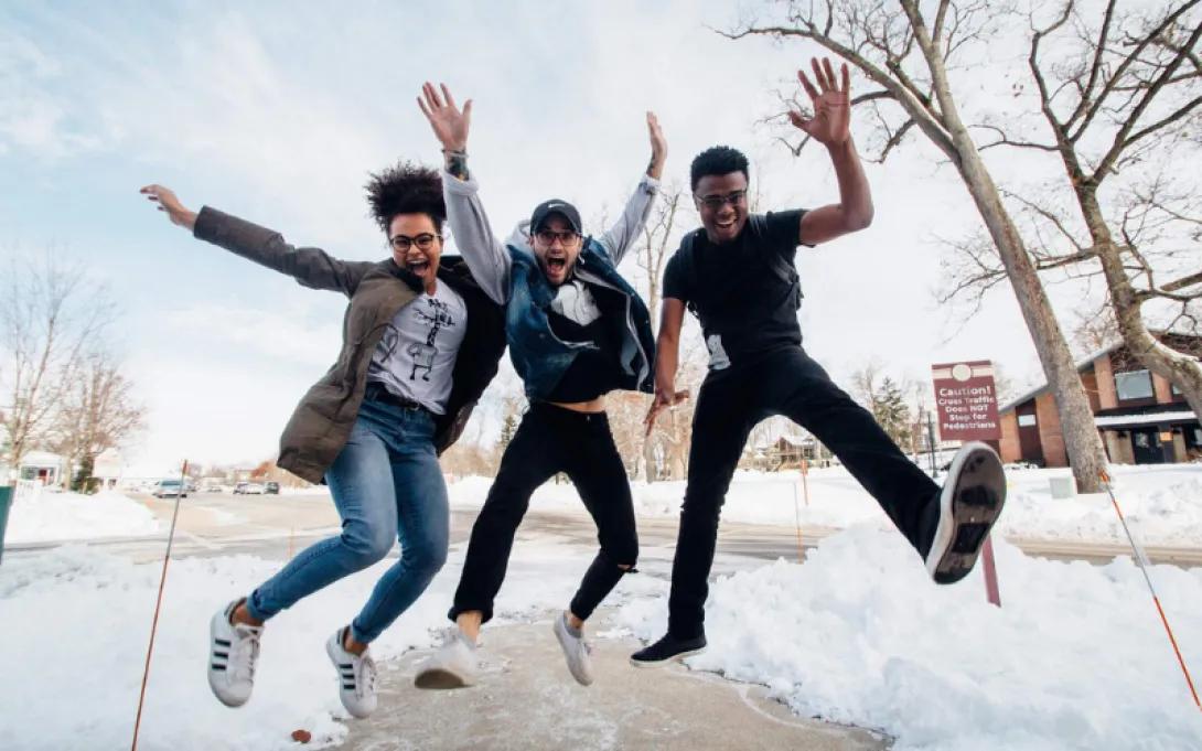 Three teens in mid-jump in an outside setting, hands raised and smiling, with snow and bare trees in the background