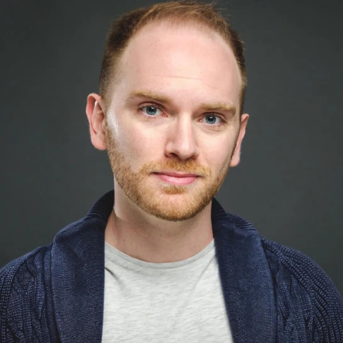 Young white man with reddish brown facial hair wearing navy blue jacket and gray crew neck shirt