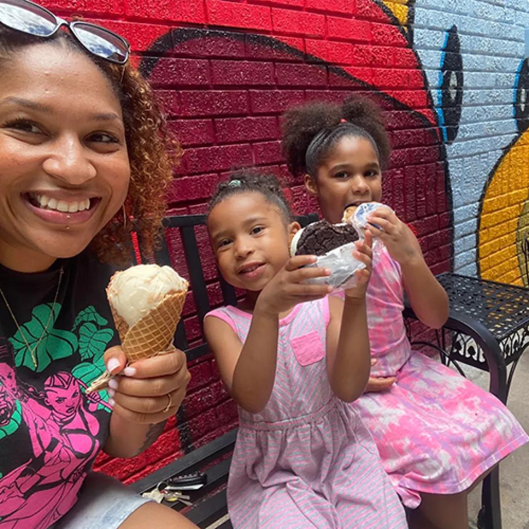 Black woman eating ice cream cone with two little Black girls wearing pink dresses