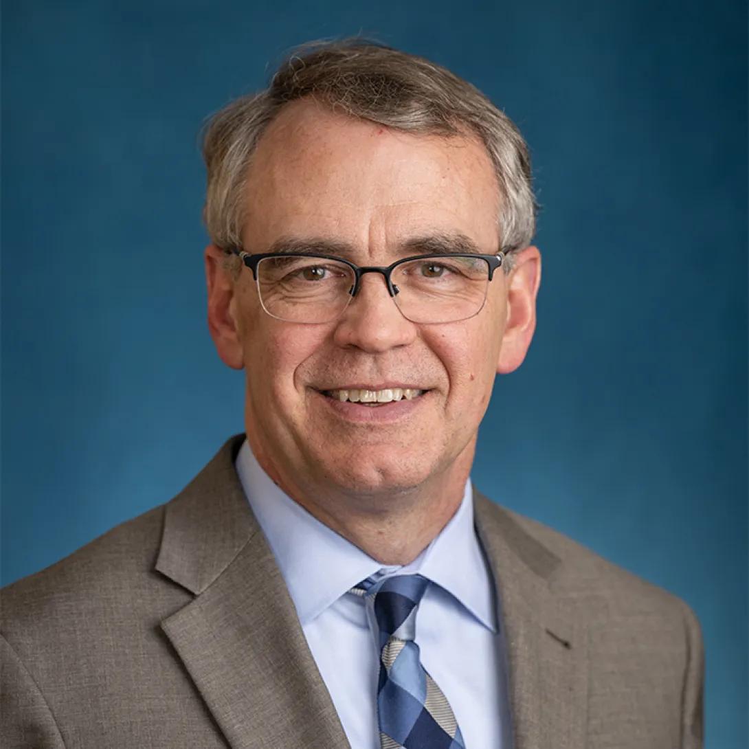Headshot of Charles Reuland - white man wearing glasses, light blue shirt and brown jacket with patterned tie