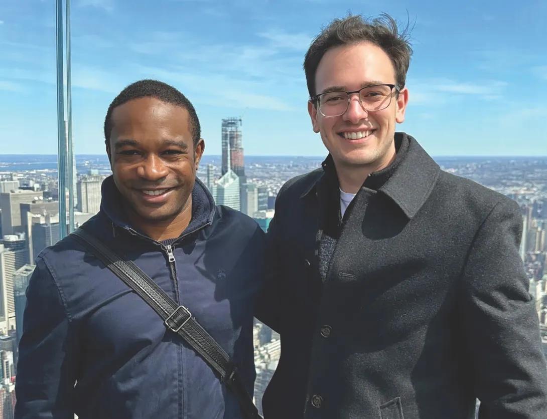 Two people standing in front of a big window with New York City in the background.