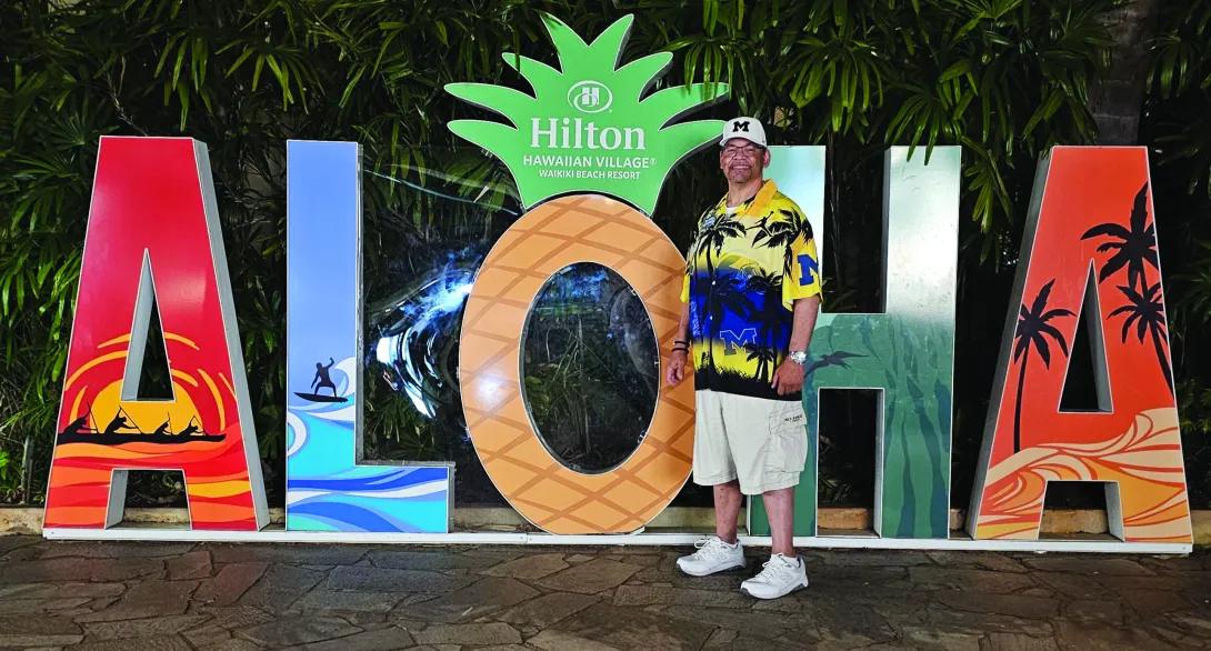 A man stands in front of a large sign that says aloha in capital letters. The letter O is in the shape of a pineapple. 