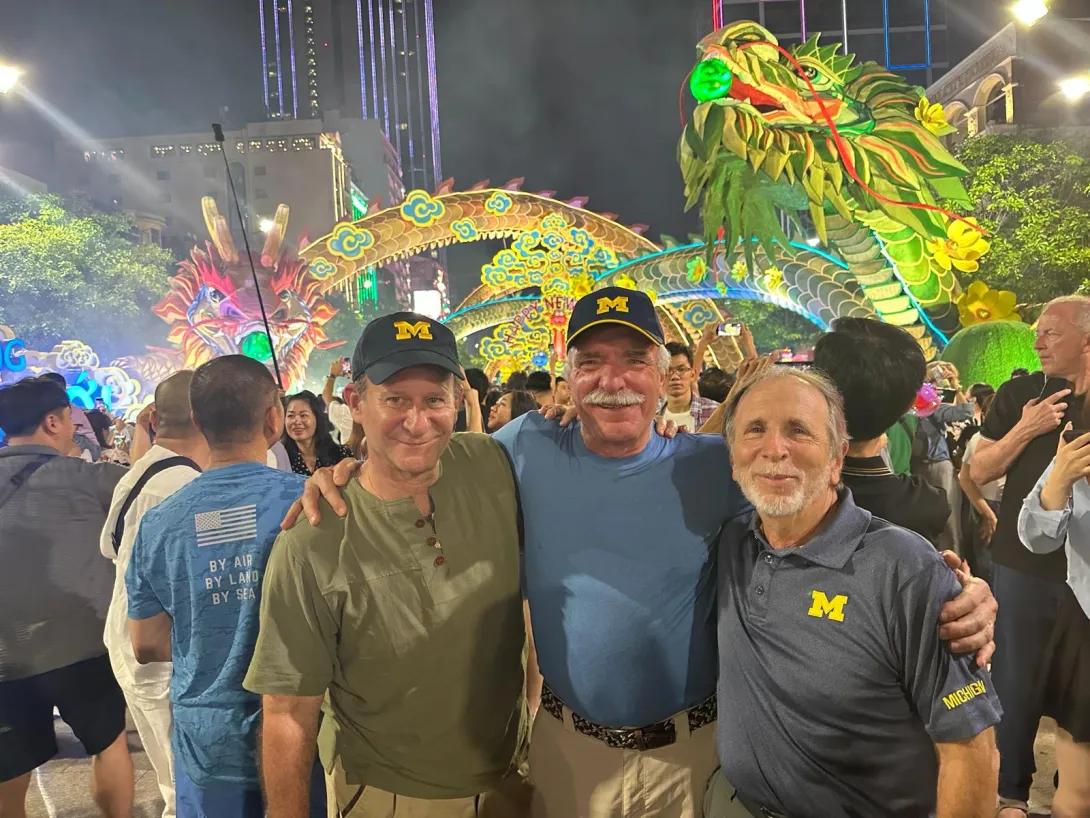 Three men are standing in a street in Vietnam with a colorful dragon in the background. The photo is taken during the Lunar New Year festival.