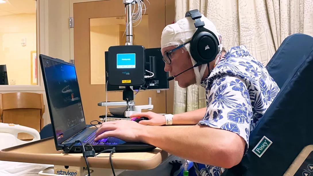 young man typing on laptop with head bandage in white on head and then headphones on ears looking at laptop screen in hospital bed