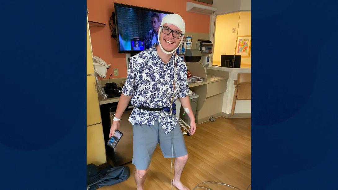 young male standing with white bandage over head smiling in patient room