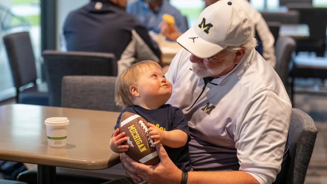 A man holding a small child on his lap. They are smiling at each other and holding a U-M football.