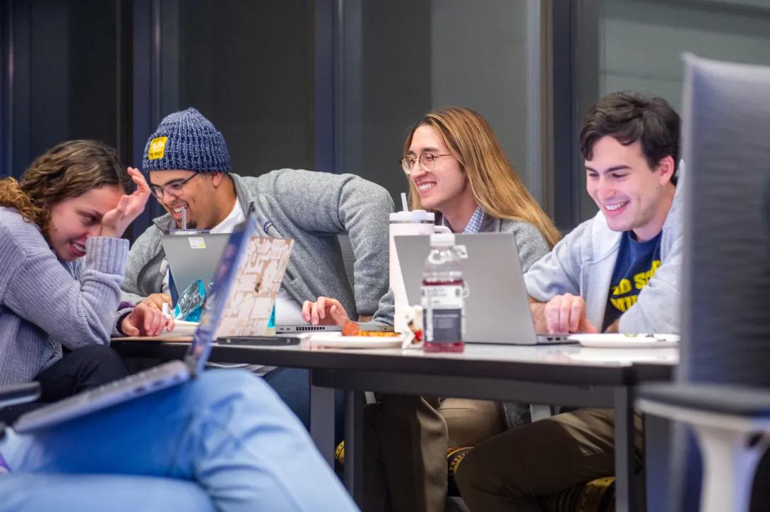 Photo by Keith-Allen Melong of students doing a read-through of the Galens Smoker annual spoof show. The students are sitting around a desk, laughing with each other. They have laptops open and beverages and snacks out on the table.