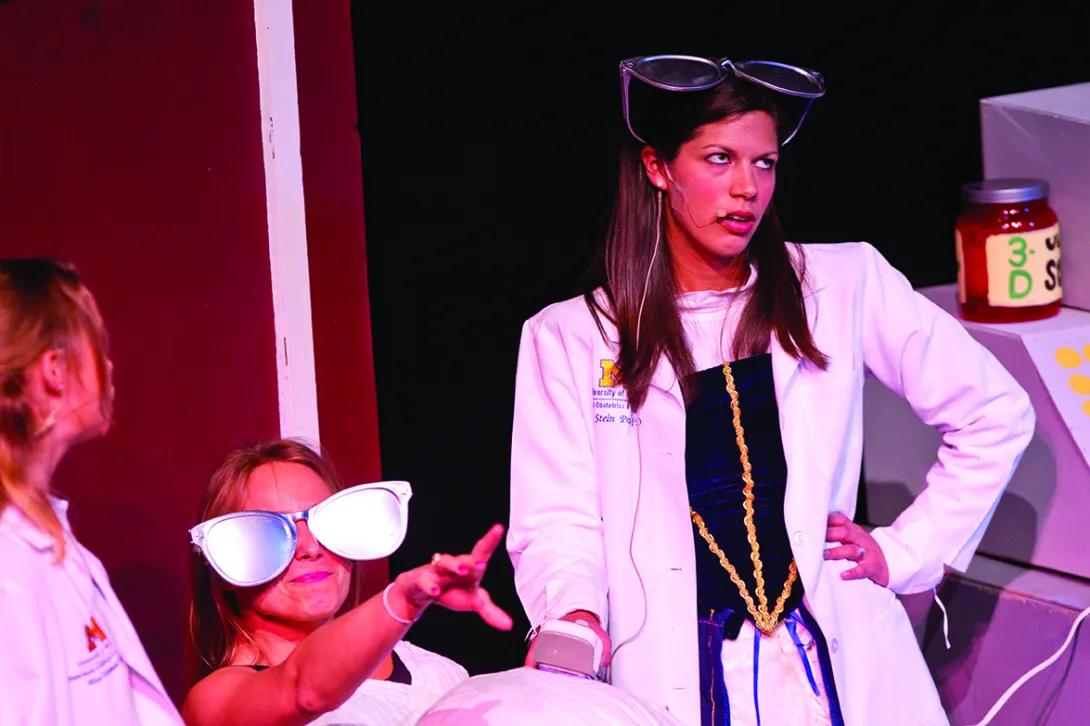 Photo of a Medical Student on stage in a costume that includes a medieval peasant dress, a doctor's white coat, and giant sunglasses on her head. She has one hand on her hip and she's rolling her eyes dramatically. Another student in giant sunglasses is kneeling next to her and pointing a finger out into the audience. They are performing in the Galens Smoker show "Thrombin Hood," a spoof of "Robin Hood."