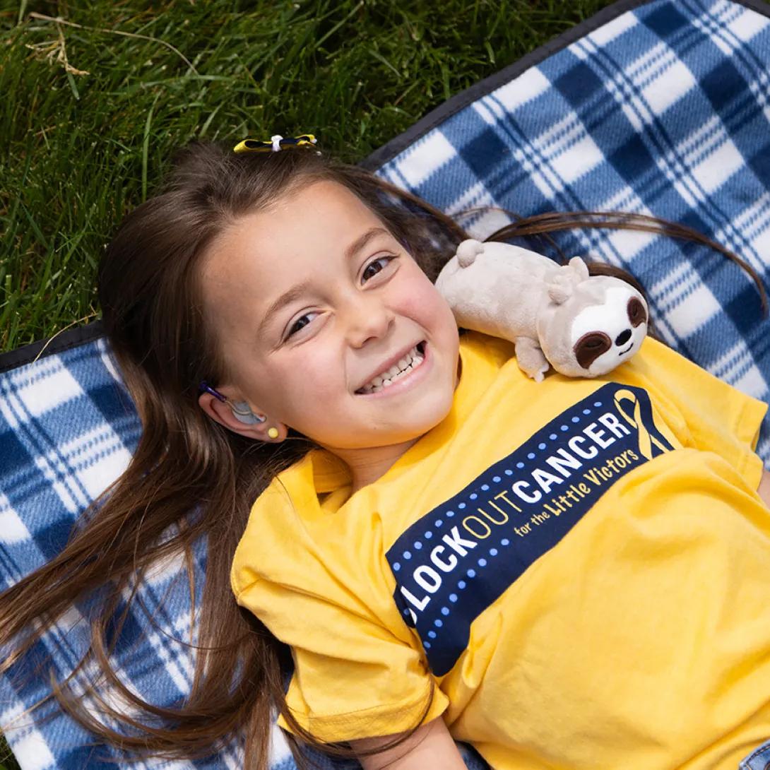 Smiling portrait of a girl in a Block Out Cancer t-shirt lying on a plaid blanket on a lawn. A plush sloth is leaned against her shoulder.