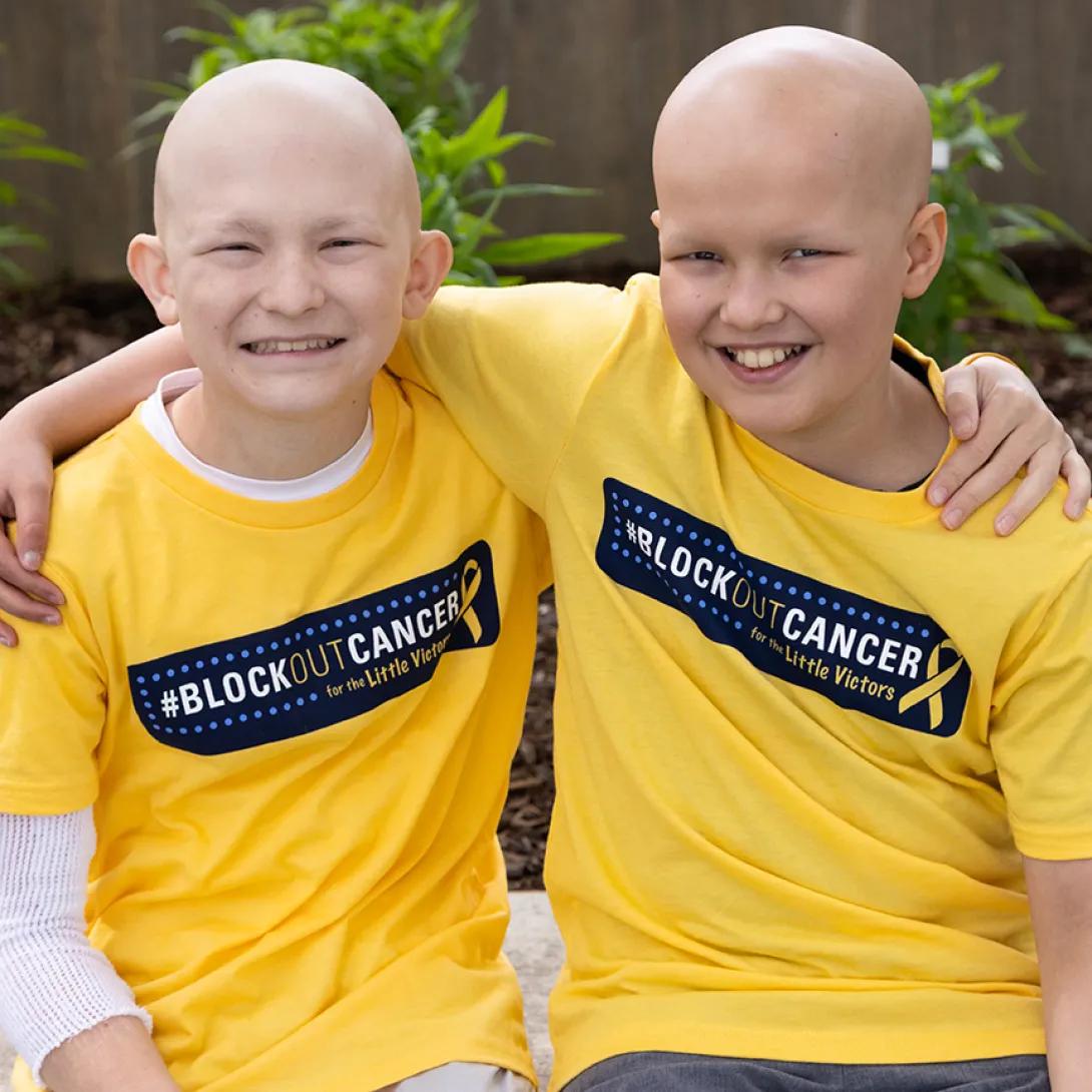Seated portrait of two boys wearing Block Out Cancer t-shirts. They are smiling and have their arms over each other's shoulders. 