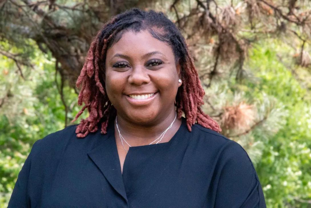 A picture of AHI's Program Specialist, Iman Saleem. Iman describes herself as having red hair, brown skin, and dark brown eyes. She wears a black top in front of a forested background.