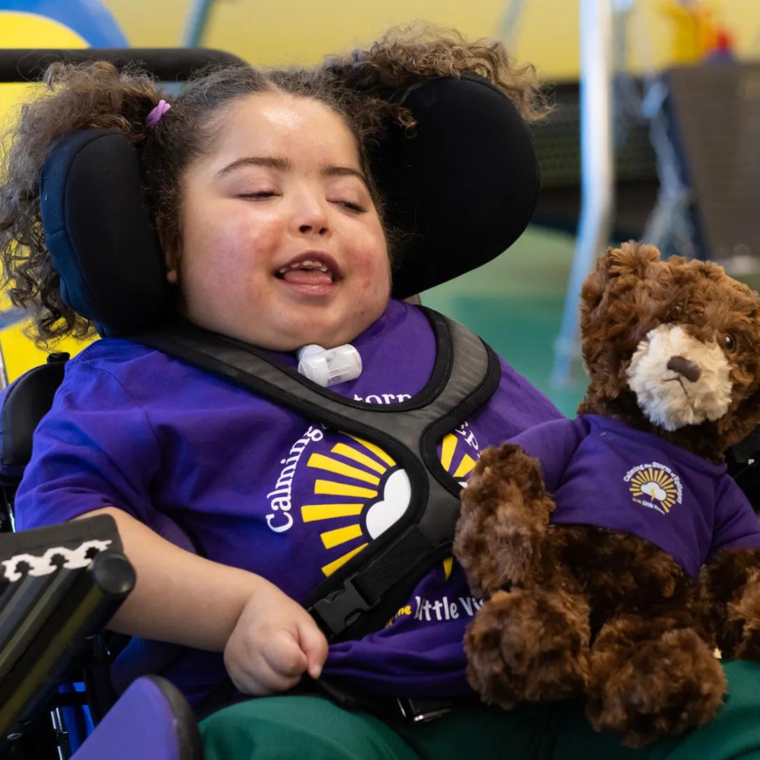 Portrait of a young girl with curly dark hair in pigtails. She is wearing a purple Calming the Storm t-shirt and is holding a brown Mott teddy bear that is wearing its own matching t-shirt.