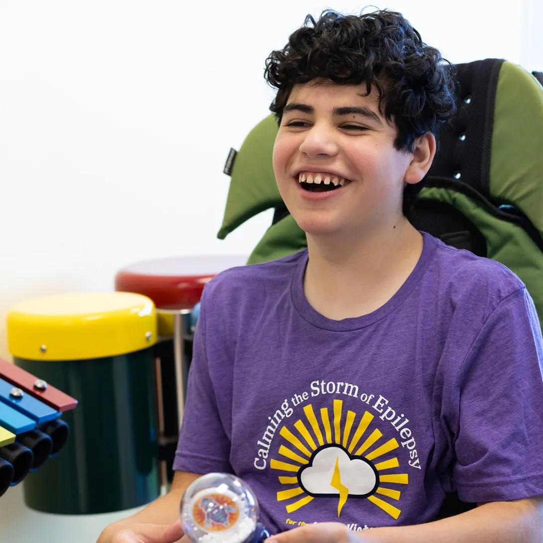 Smiling portrait of a smiling boy with dark curly hair. He is wearing a purple Calming the Storm t-shirt. A xylophone and two percussion instruments are visible to the side of him.