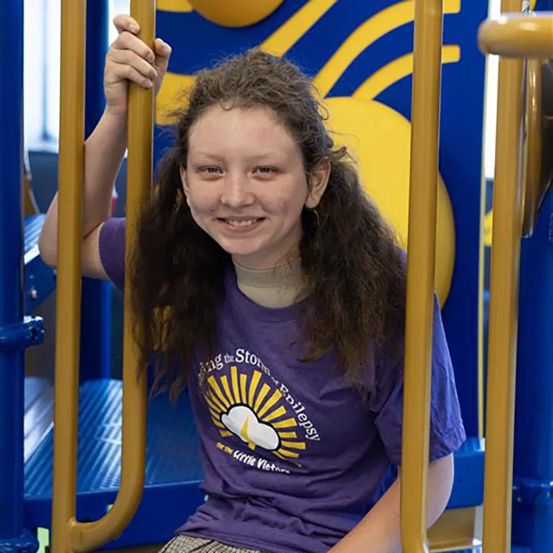 Seated portrait of a girl with long, brown, wavy hair. She is wearing a purple Calming the Storm t-shirt and appears to be seated on play equipment.