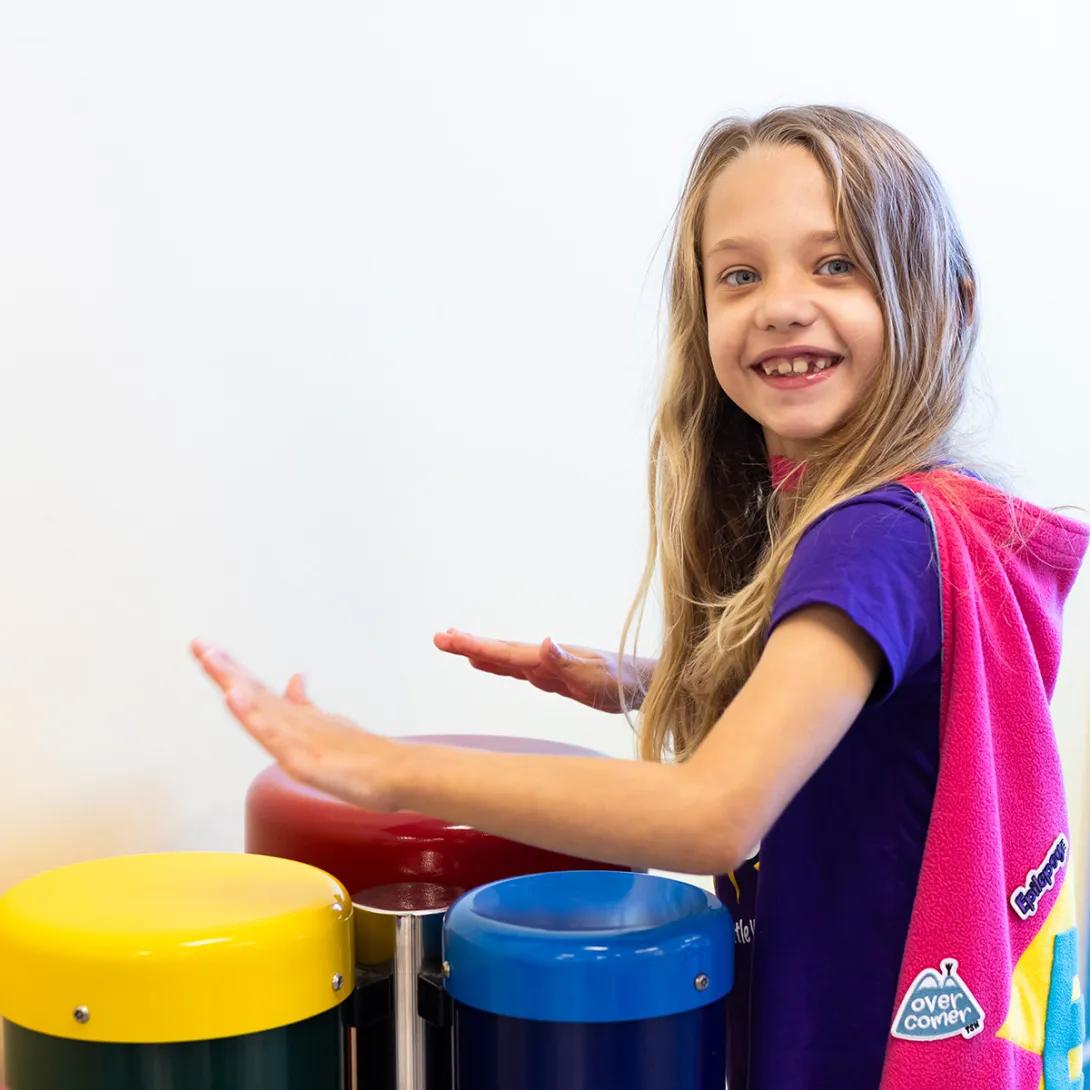 Evalynn playing three toy percussion instruments that look like drums. Evalynn is facing to the left but is smiling and has her face turned toward the viewer. She is wearing a purple Calming the Storm t-shirt and a pink superhero cape.
