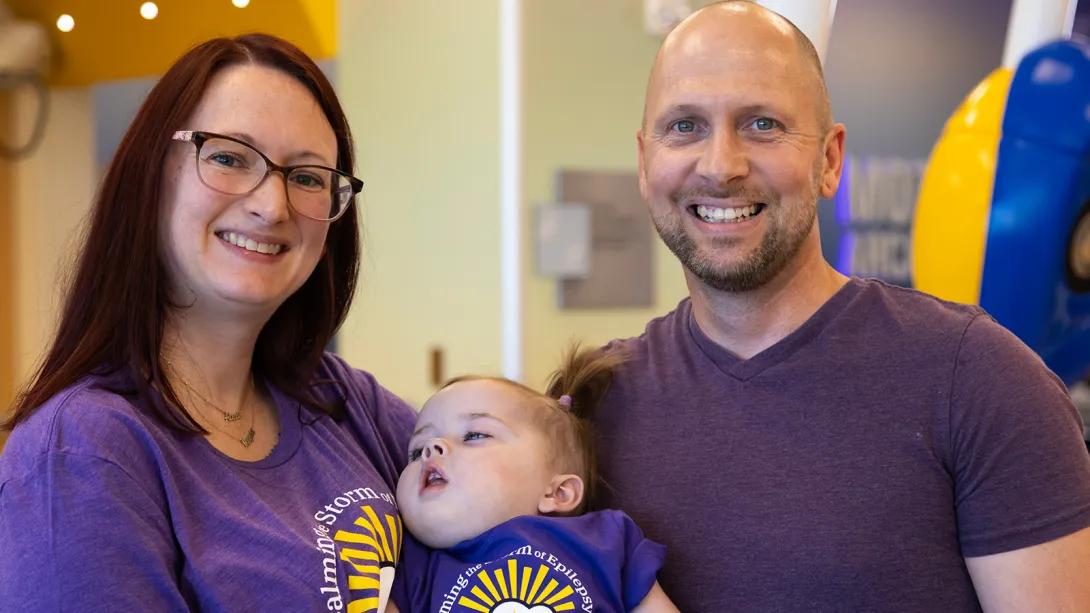 Smiling portrait of two adults, one of whom is wearing a purple Calming the Storm t-shirt. They are holding an infant between them who is also wearing a purple Calming the Storm t-shirt.