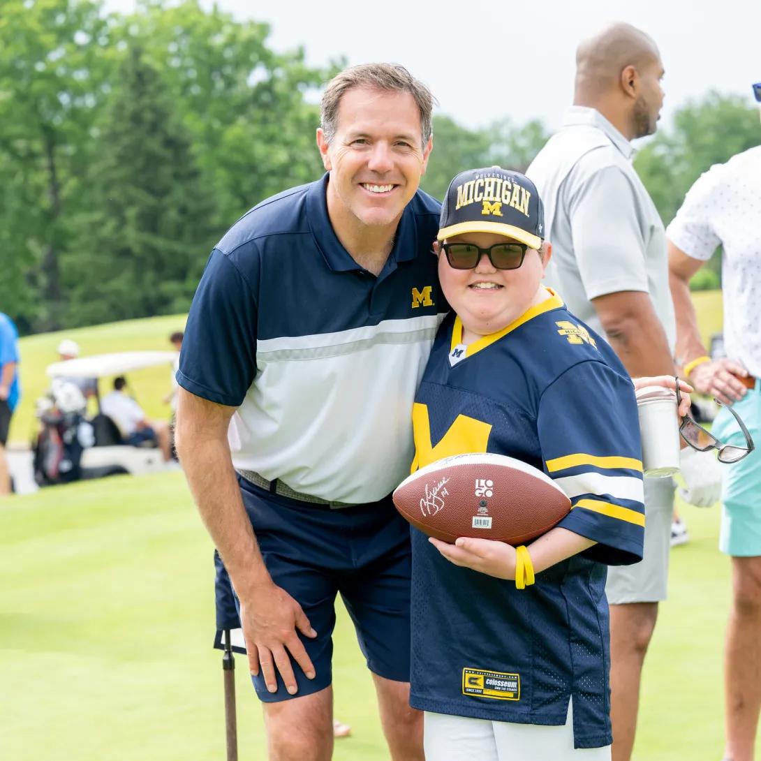Two people wearing U-M gear lean in towards each other and smile at the viewer. One is holding a football. They are on the course at the Golf on Main event and a few golfers and a cart are visible behind them. 