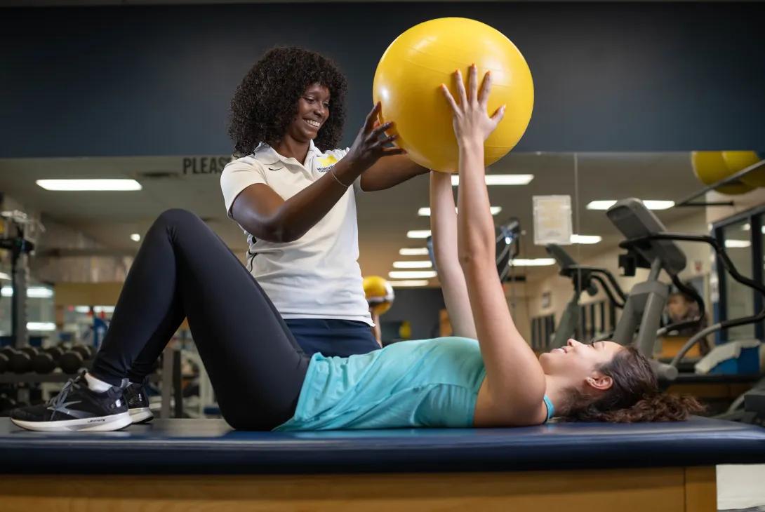 A sports medicine professional stands next to an athlete who is lying on a table in a gym. They are both holding a yellow ball up to guide the athlete in an exercise.