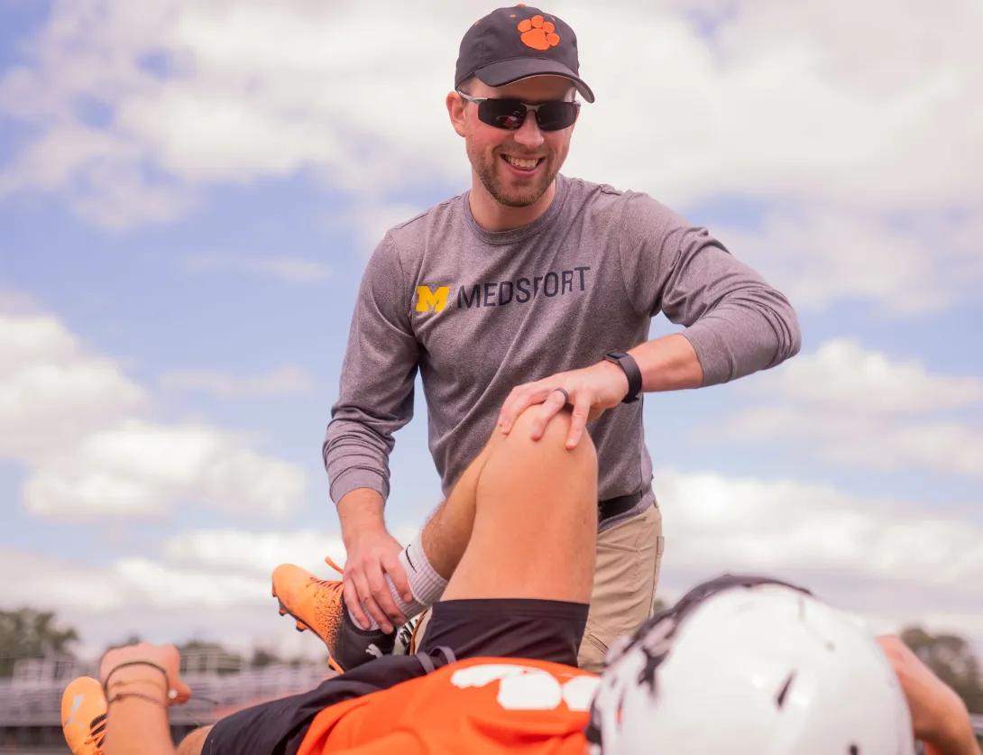 A high school football player in an orange jersey is lying on a football field and a sports medicine professional in a U-M med sport t-shirt kneels next to him to help him stretch.