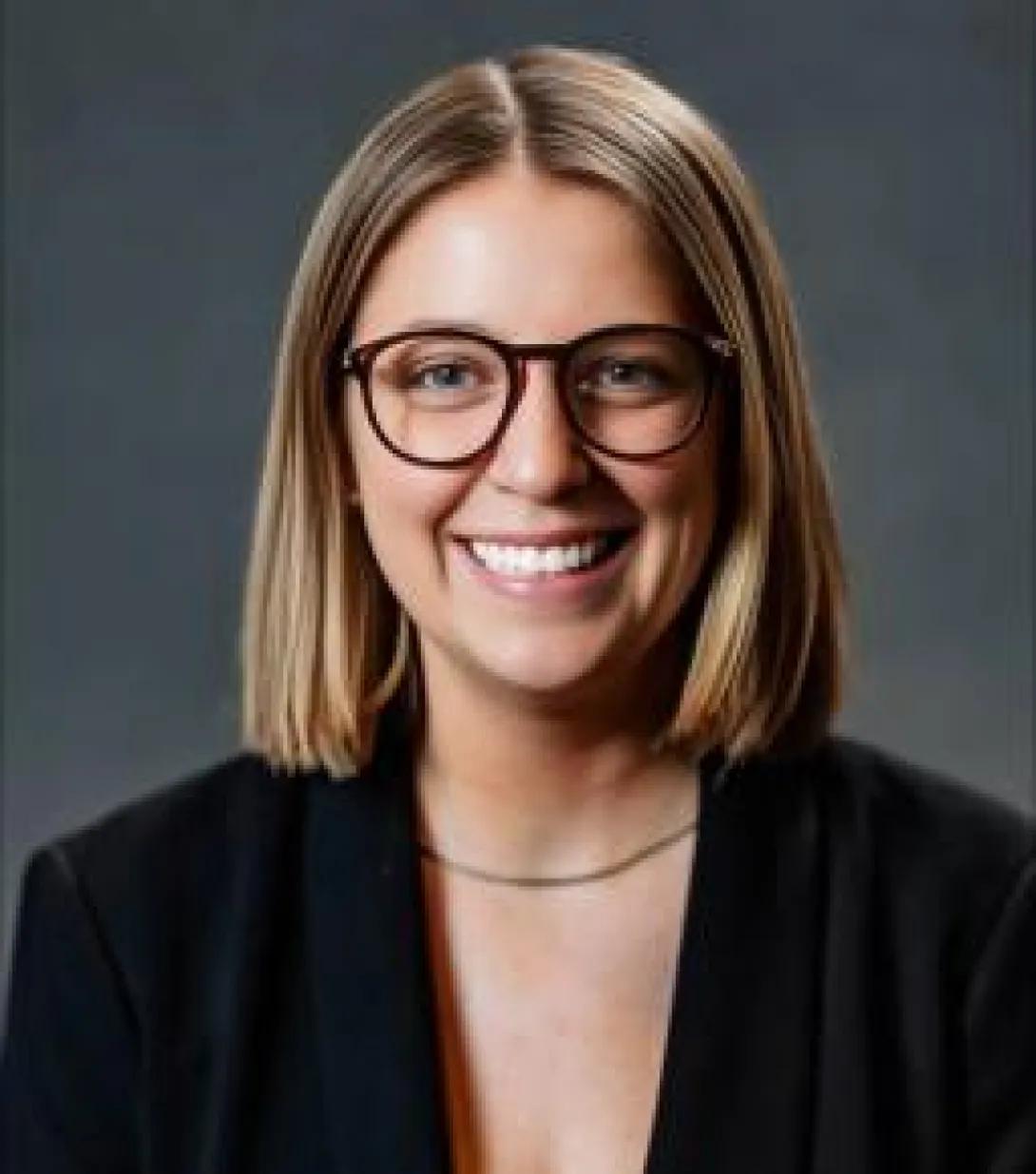 Smiling portrait of Shae Maier. She is wearing glasses, a dark blazer, and a simple necklace.