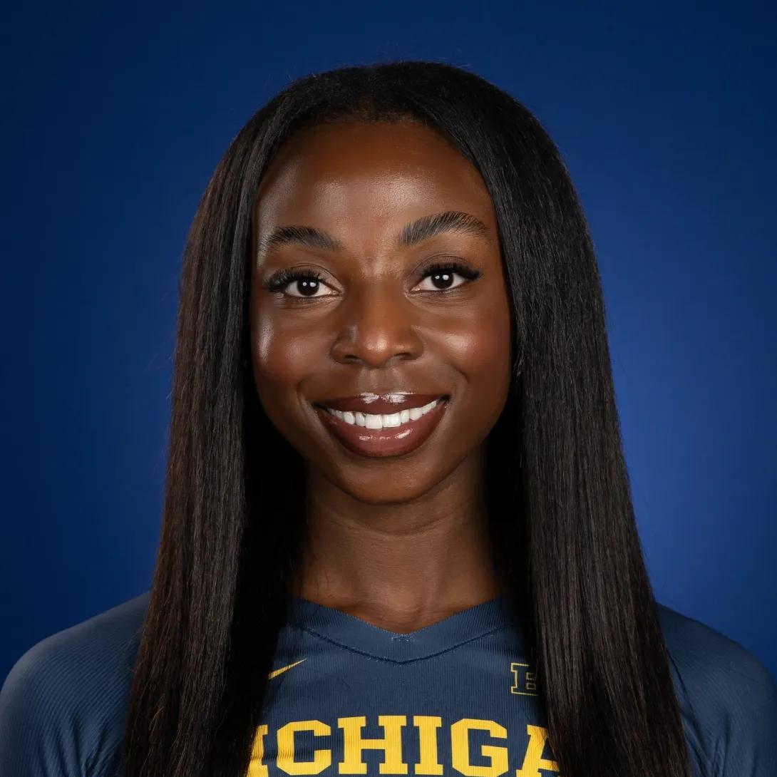 Jacyn Bamis, smiling in front of a blue background wearing a Michigan sports jersey. Jacyn describes herself as having dark brown eyes, brown skin, and dark black hair.