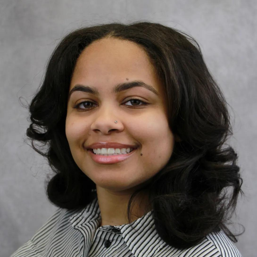 Jordan Bell, smiling and wearing a striped shirt in front of a grey background. Jordan describes herself as having beige/honey skin, sandy brown hair, and hazel eyes.