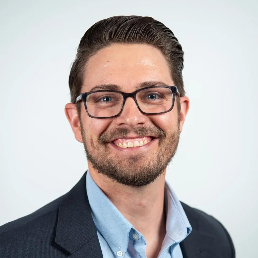 Smiling headshot of Ryann Eff. He is wearing glasses and dark blue jacket with a light blue button-down.